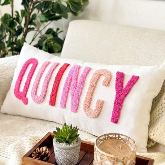 White pillow with 'QUINCY' in pink letters on a couch with a plant and decor items in the background.