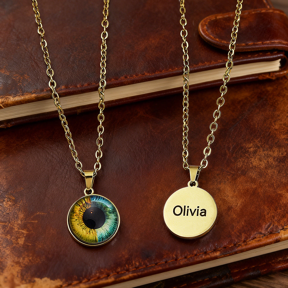 Two gold necklaces on a leather surface, one with an eye design pendant and the other with a 'Olivia' engraved pendant.