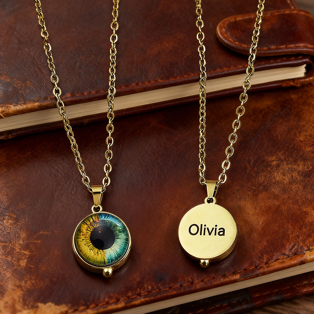 Two gold necklaces on a leather surface, one with an eye design and the other with 'Olivia' engraved.