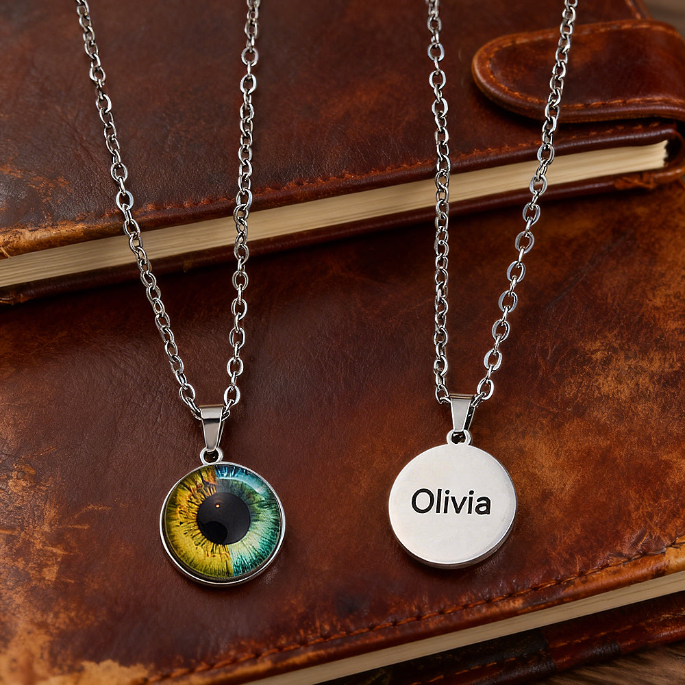 Two silver necklaces on a brown leather surface, one with an eye design and the other with 'Olivia' engraved.