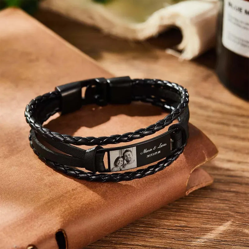 Black braided leather bracelet with engraved tag on a brown leather surface